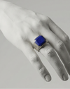 Hand wearing a ring with a blue gemstone on a neutral background