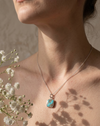 Necklace with a turquoise pendant worn by a person, with white flowers in the background.