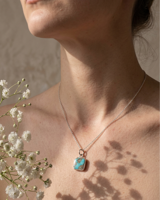Necklace with a turquoise pendant worn by a person, with white flowers in the background.
