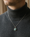Man wearing a dark turtleneck sweater with a necklace featuring a square pendant.