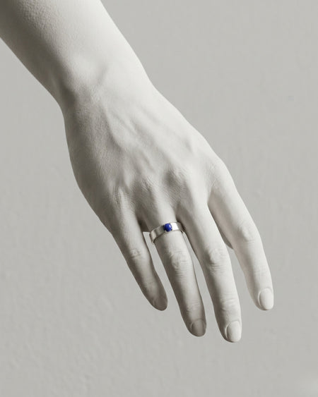White hand wearing a ring with a blue gemstone on a light gray background