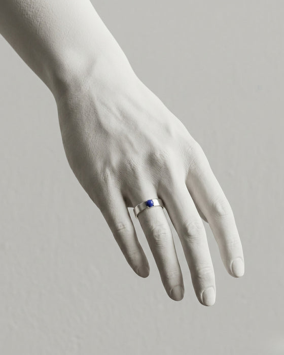 White hand wearing a ring with a blue gemstone on a light gray background