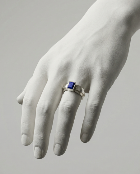 White marble hand wearing a silver ring with a blue gemstone on a gray background